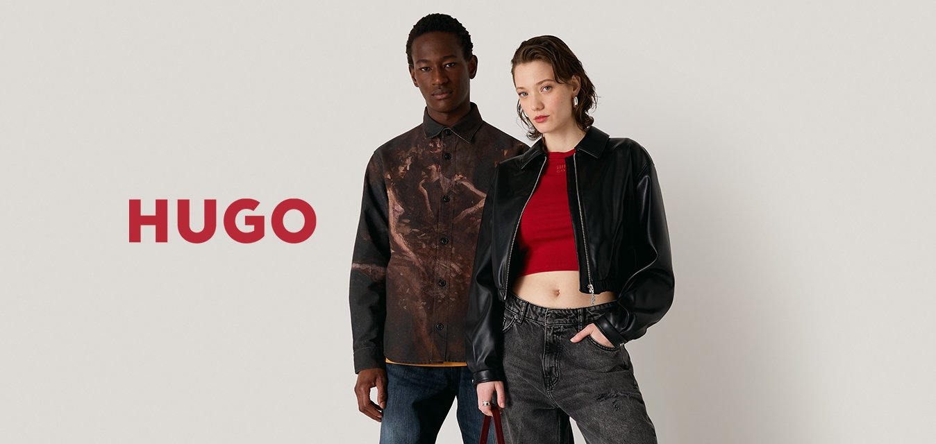 Dvě modelky stojí na neutrálním pozadí. Jedna má na sobě vzorovanou hnědou košili a modré džíny, druhá černou zkrácenou bundu a šedé kalhoty.