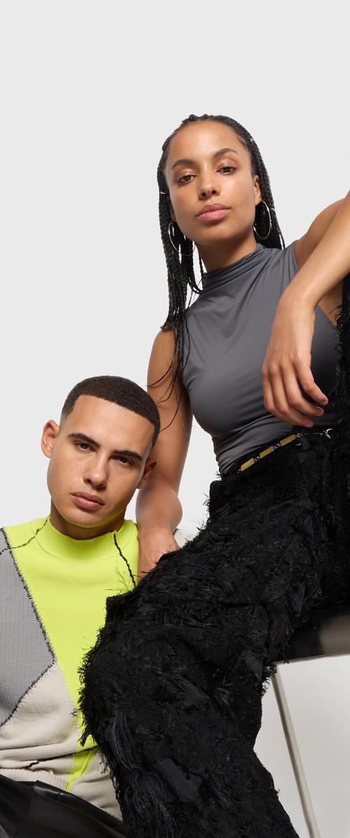 Deux individus posent ensemble. L'un porte un haut gris ajusté et un pantalon noir texturé, tandis que l'autre arbore un pull vert néon avec des motifs géométriques.