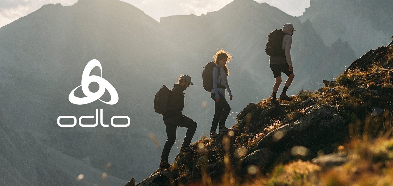 Tři turisté s batohy stoupající po kamenitém svahu hory pod zataženou oblohou s vzdálenými vrcholky na pozadí.