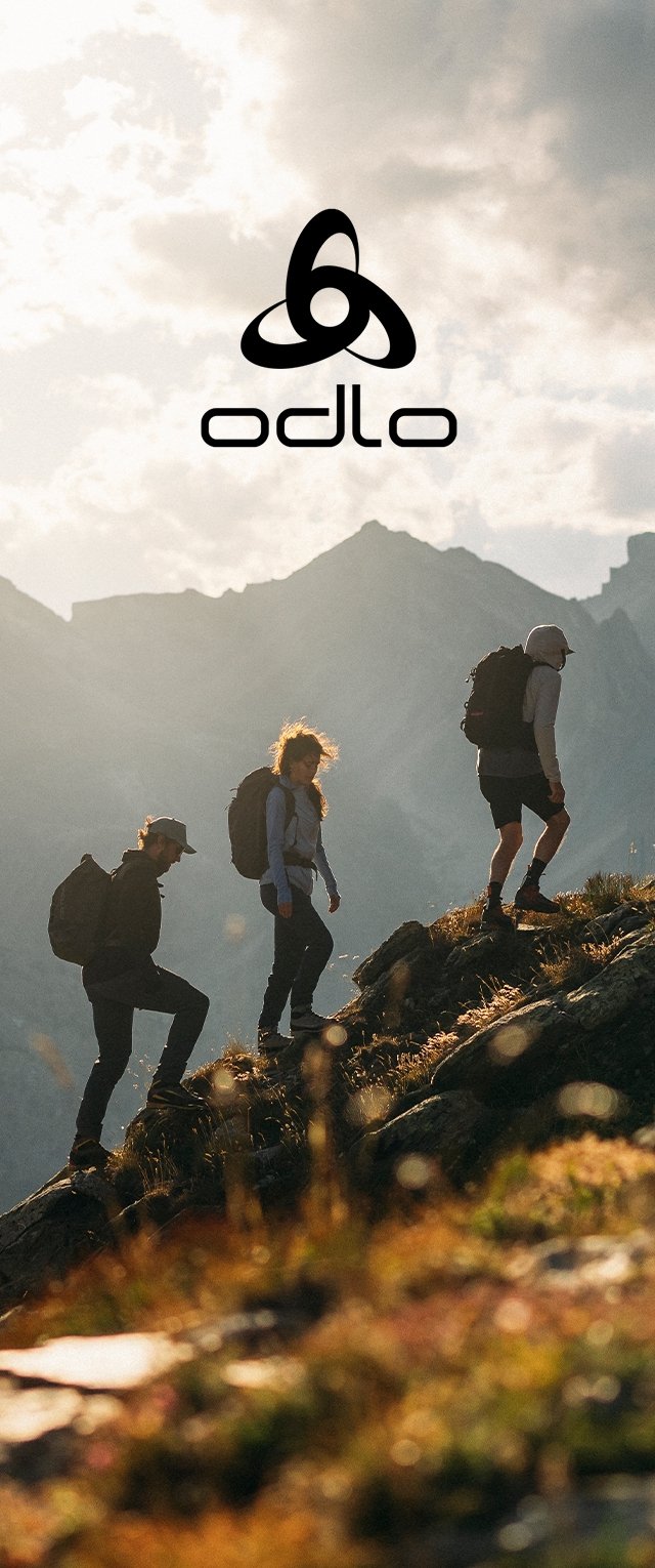 Drei Wanderer steigen einen steinigen Hang hinauf, während sie Rucksäcke tragen. Die Landschaft zeigt zerklüftete Berge und goldenes Gras unter einem dramatischen Himmel.