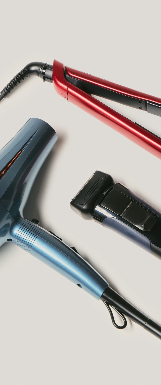 Tres herramientas para el peinado: un secador de cabello azul, una plancha para el pelo roja y una afeitadora eléctrica negra con azul, con diseños de plástico elegantes y cables.