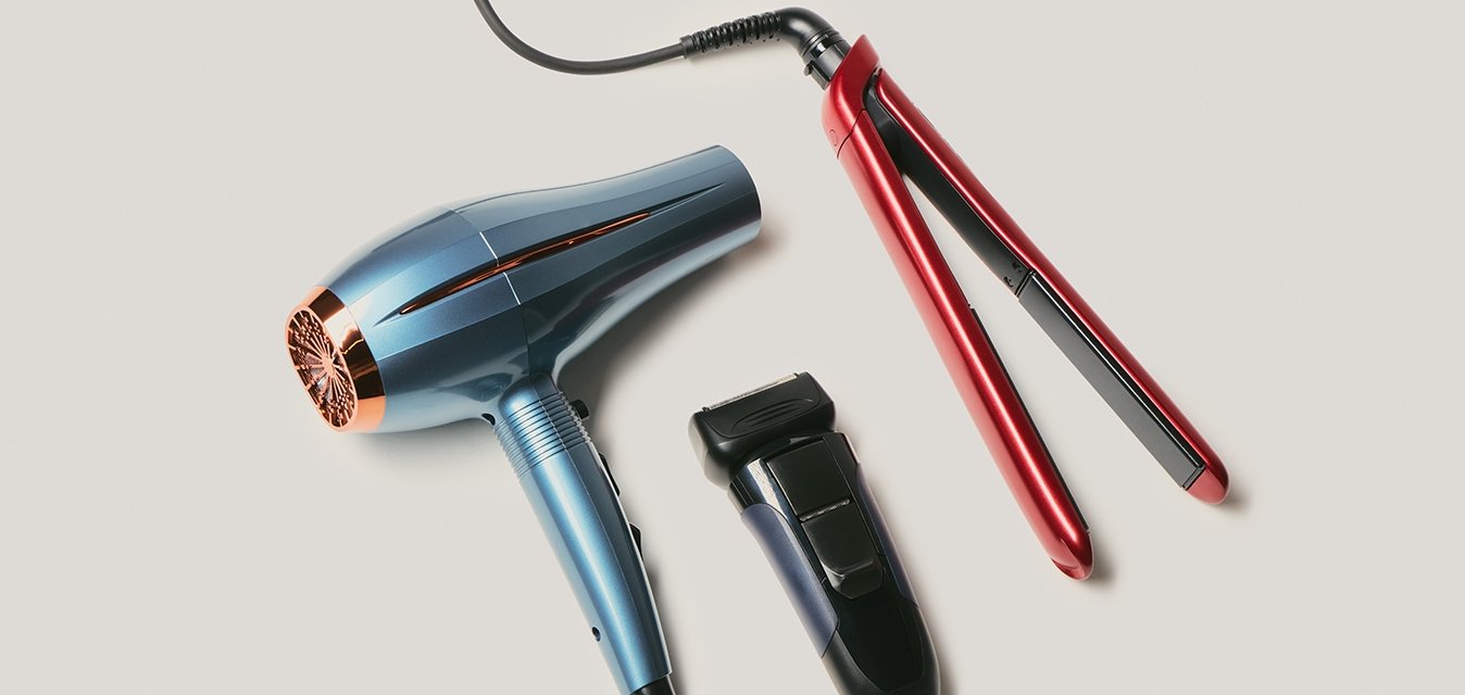 Modrý vysoušeč vlasů, červená žehlička na vlasy s kabelem a černý elektrický holicí strojek uspořádané na světle béžovém povrchu.