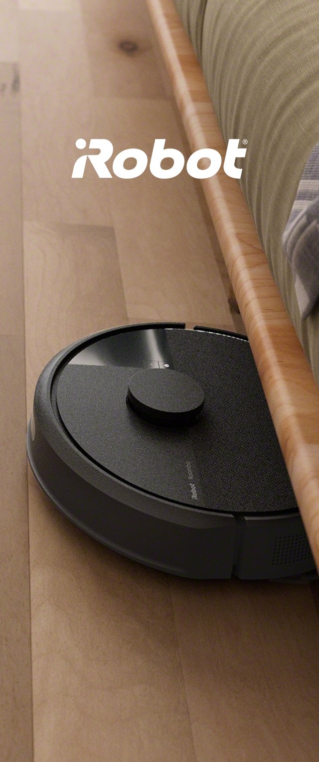 Un robot aspirador negro y redondo reposa contra un suelo de madera, parcialmente escondido bajo una cama. Presenta una superficie texturizada y un botón central prominente.