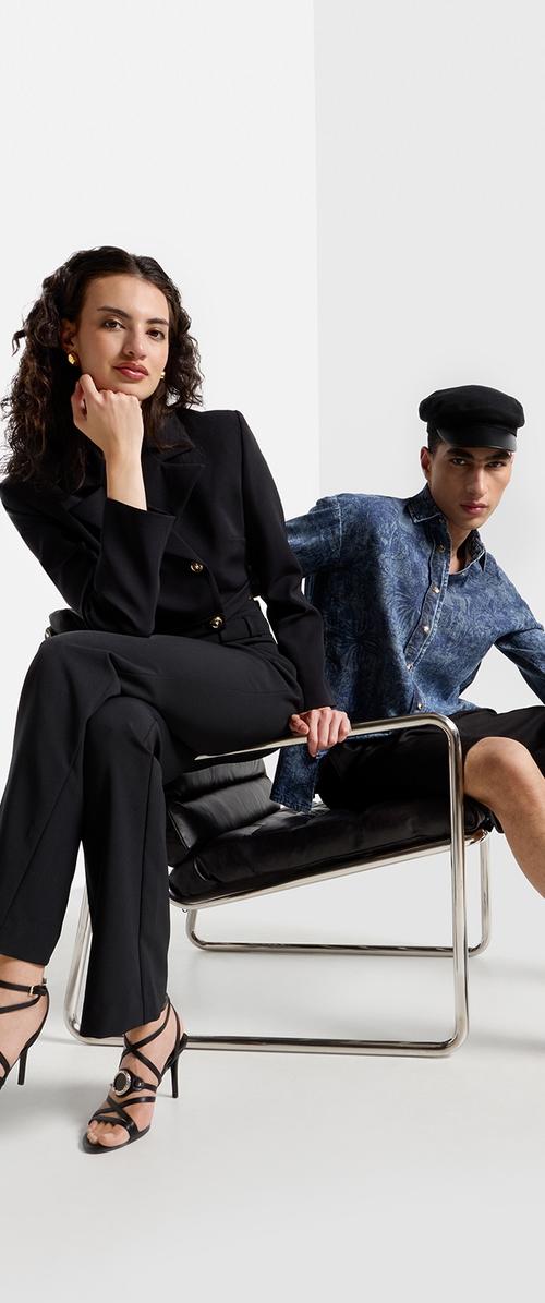 Dos modelos sentados en una silla de cuero negro. Uno lleva un traje negro con botones dorados; el otro tiene una camisa de patrón azul y una gorra negra.