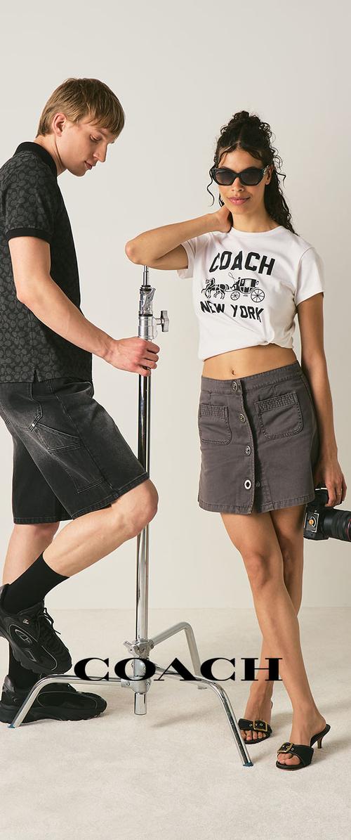 Een man in zwarte short en hemd houdt een metalen standaard vast terwijl een vrouw met een zonnebril, een wit cropped Coach New York shirt en een grijze rok poseert.