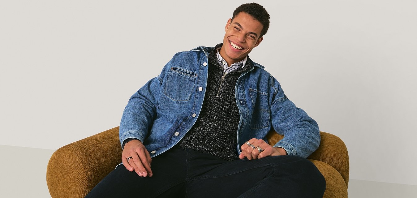 Mladý muž se usmívá, pohodlně sedí na hnědém texturovaném křesle na jednoduchém světlém pozadí.