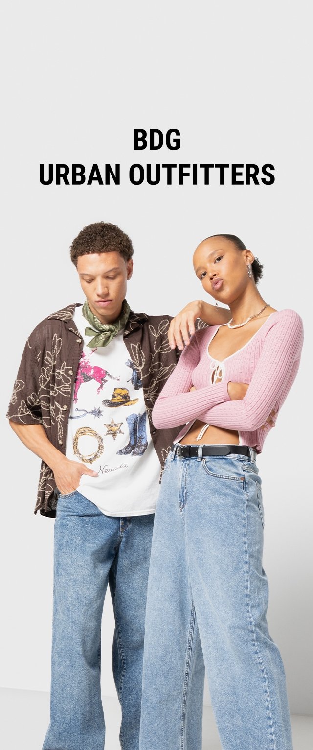Dos personas posan vistiendo ropa casual. Uno lleva una camisa de manga corta marrón con estampado, una camiseta gráfica y unos vaqueros de mezclilla azules. El otro lleva un top de canalé rosa, vaqueros azules y accesorios como un collar.