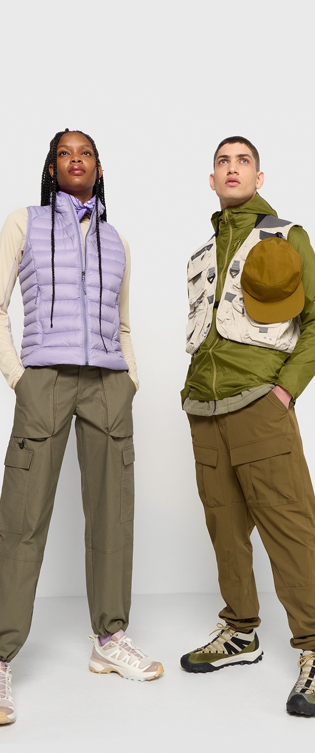 Ljuslila puffväst, grön lagerjacka, cargobyxor och beige sneakers med flerfärgade accenter, bärs av två modeller mot en vit bakgrund.