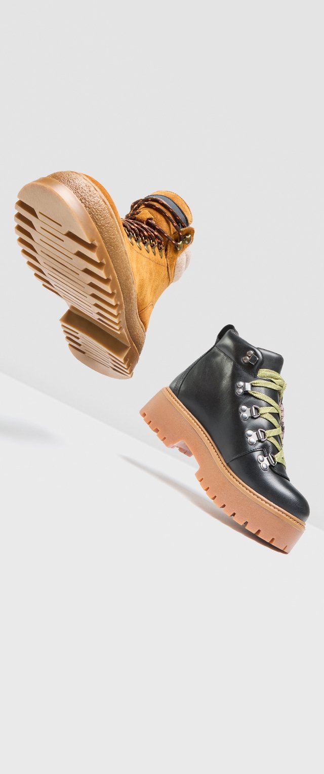 Braune und schwarze Stiefel mit Gummisohlen. Der braune Stiefel hat Schnürsenkel und eine weiche Textur, während der schwarze Stiefel metallische Akzente und grüne Schnürsenkel aufweist.