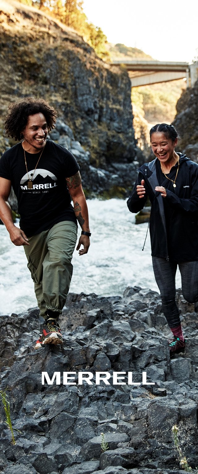 Deux personnes marchent sur un terrain rocailleux à côté d'une rivière. L'une porte un t-shirt noir et un pantalon cargo, l'autre une veste noire et des leggings à motifs.