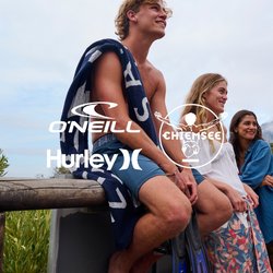 Tři jednotlivci sedí na dřevěném zábradlí, oblečeni v neformálním oblečení. Krajina zahrnuje zeleň, skalnaté kopce a zataženou oblohu.