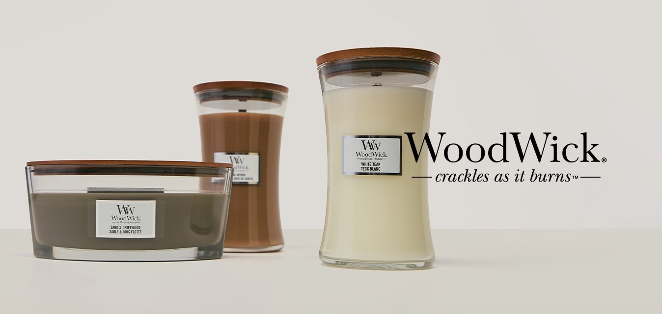 Tři svíčky WoodWick s dřevěnými víčky, v oválných a přesýpacích skleněných nádobách, uspořádané na hladkém bílém povrchu.