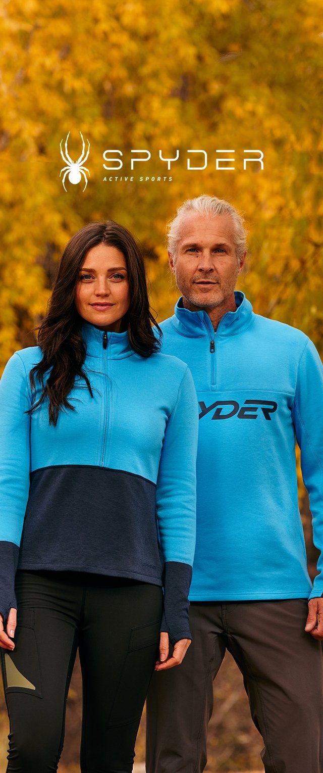 Dos adultos vestidos con chaquetas deportivas Spyder de color azul y negro se encuentran al aire libre frente a árboles de otoño con hojas amarillas.