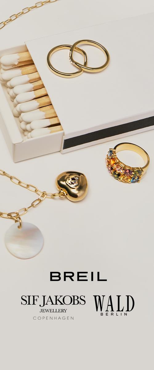 Zwei Goldringe auf einer Streichholzschachtel, ein Goldring mit mehrfarbigen Steinen und eine Goldkette mit Herz- und Scheibenanhängern auf weißer Oberfläche.