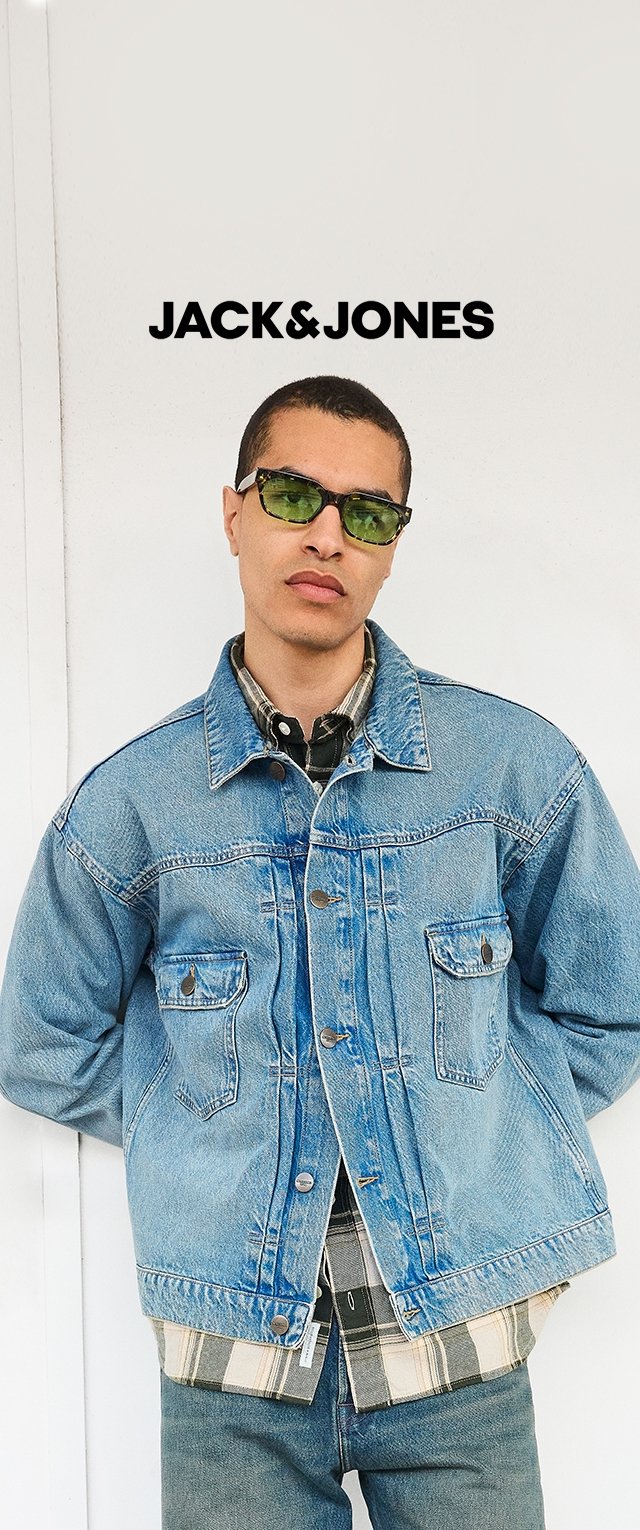 Jeune homme portant des lunettes de soleil vertes, veste en jean superposée sur une chemise à carreaux, debout contre un mur blanc avec le logo "JACK & JONES" au-dessus.