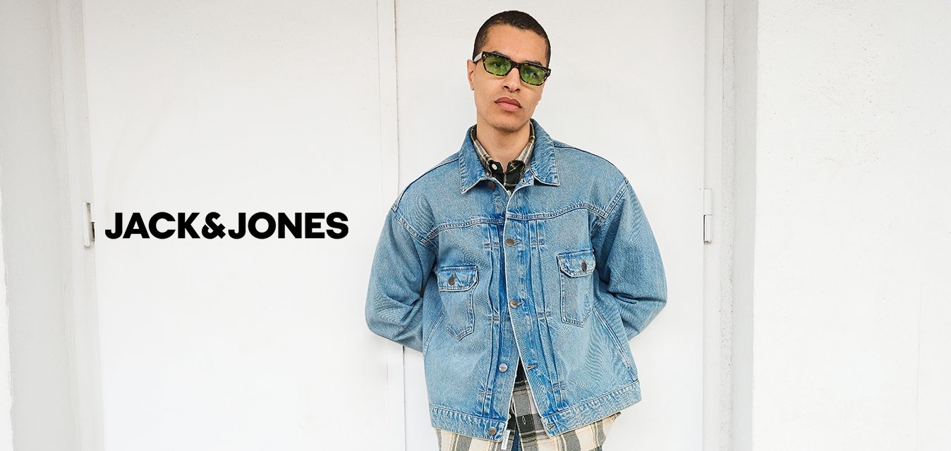 Muž s zelenými slunečními brýlemi, světlou džínovou bundou přes kostkovanou košili, modrými džíny a černými botami stojí opřený o bílou zeď s rukama za zády.