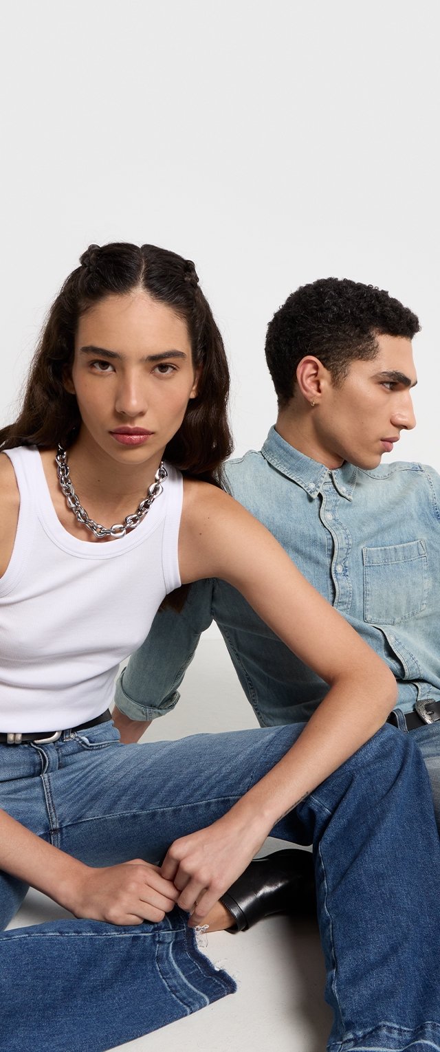 Witte mouwloze top, zilveren ketting, blauwe spijkerbroek. Eén persoon met donker haar, een ander met krullend haar en een lichtblauwe shirt.