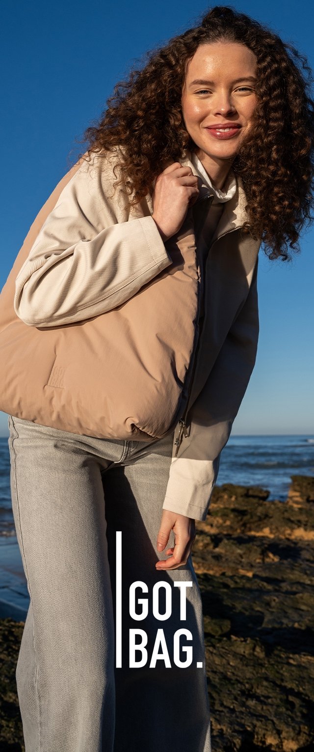 Mujer sonriente con cabello rizado lleva un gran bolso beige al hombro, parada cerca de una costa rocosa bajo un cielo azul despejado.