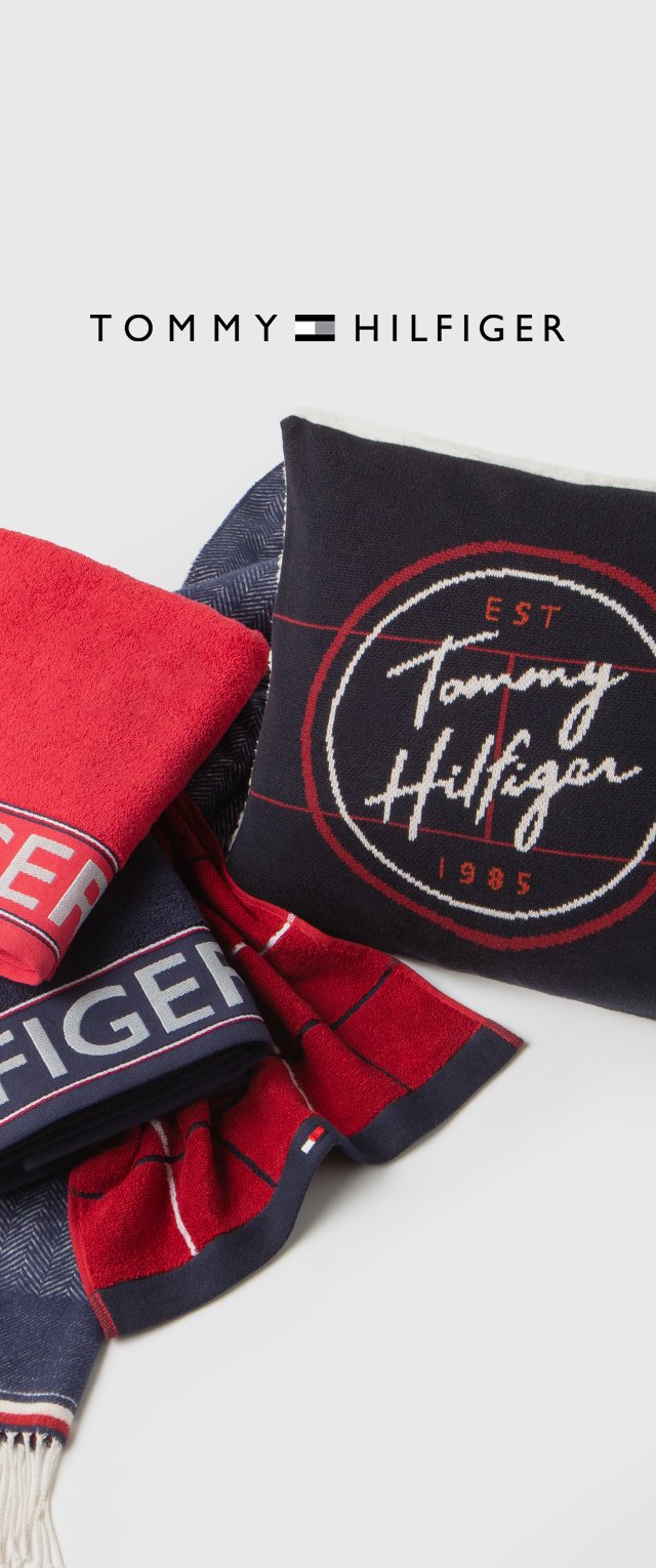 Tommy Hilfiger handdoek- en kussen set in rood en marineblauw. Handdoek heeft een gedurfde logo band; kussen heeft een cirkelvormig logo met "EST 1985." Zachte texturen.