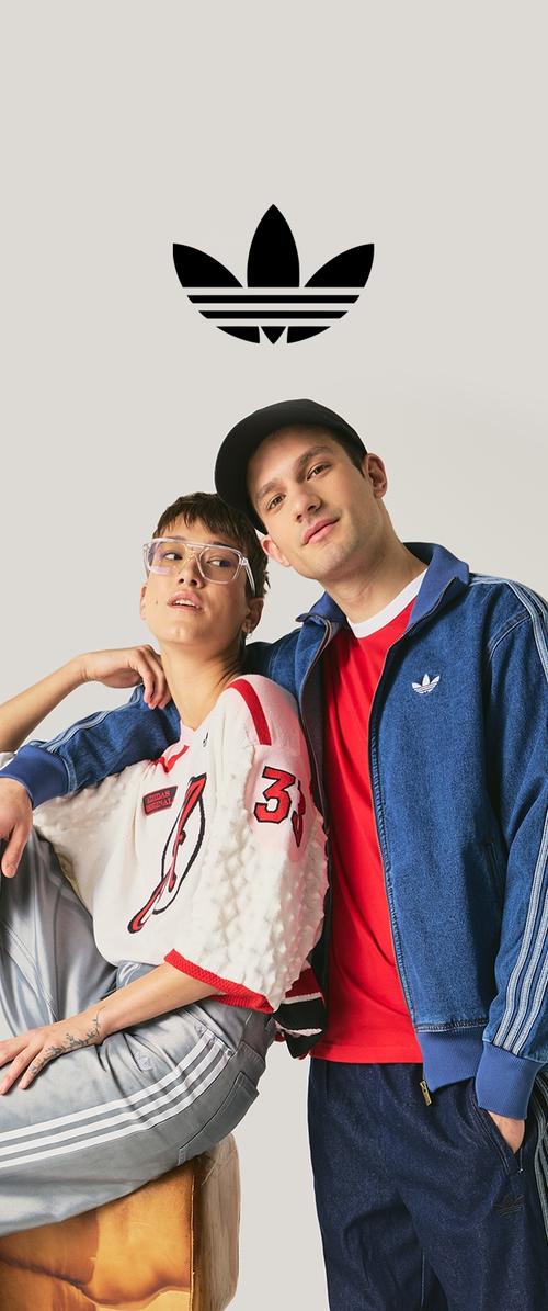 Deux jeunes adultes en tenue de sport Adidas posent sous un grand logo trefoil d'Adidas sur un fond beige.
