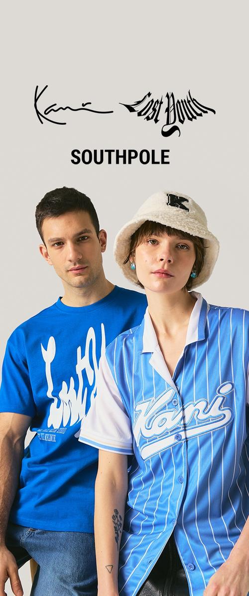 Joven con camiseta gráfica azul y joven con camisa azul de rayas y sombrero blanco de felpa con logo "K", posando frente a un fondo beige.