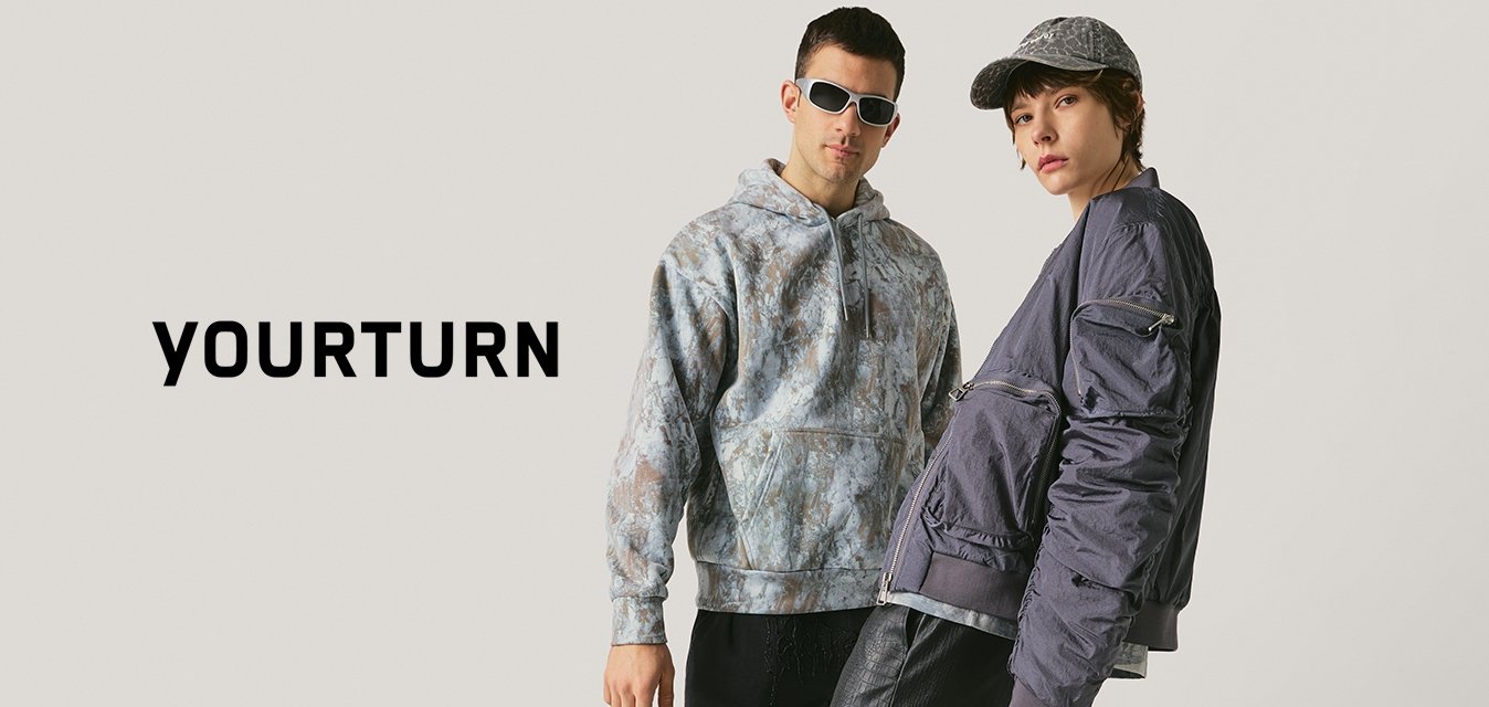 Dva módně oblečení lidé pózují před neutrálním pozadím, mají na sobě tmavé kalhoty, vrstvené topy a doplňky včetně slunečních brýlí a tašky.