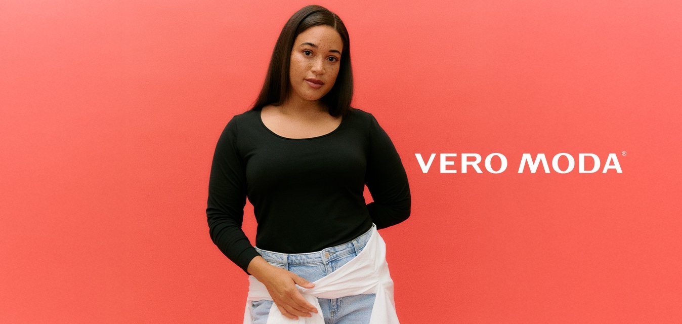 Žena s dlouhými tmavými vlasy, oblečená v černém tričku s dlouhým rukávem, světlých modrých džínách a bílou košilí uvázanou kolem pasu, na korálovém pozadí.