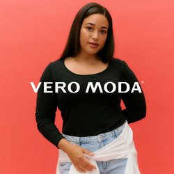 Žena s dlouhými tmavými vlasy, oblečená v černém tričku s dlouhým rukávem, světlých modrých džínách a bílou košilí uvázanou kolem pasu, na korálovém pozadí.