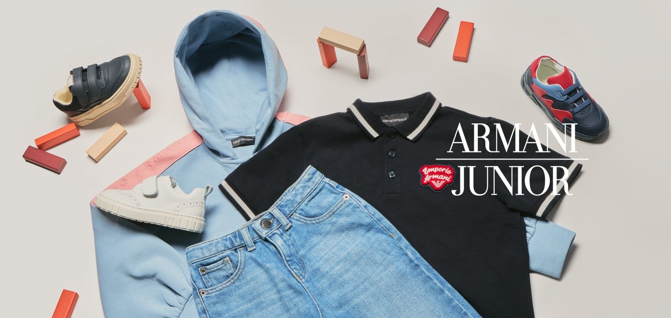 Světle modrá mikina s kapucí, černé polo tričko s červeným logem, modré džíny a tři páry barevných dětských tenisek rozložených kolem oblečení na bílém povrchu.