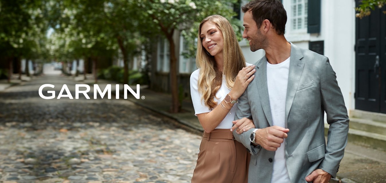 Pár kráčí po dlažební ulici. Žena v bílém svetru a béžové sukni, muž v šedém saku přes bílém tričku, stromy lemují cestu.