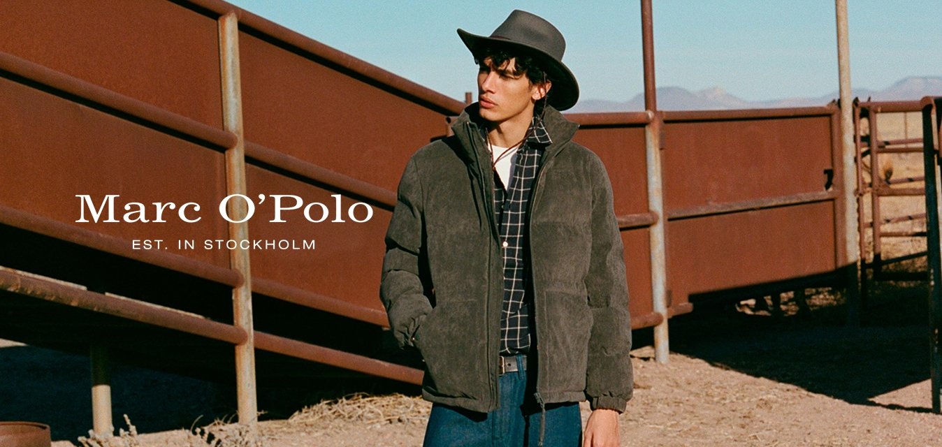 Muž v tmavé bundě přes kostkovanou košili a modré džíny stojí poblíž kovových plotů, v pozadí rezavý sud a pouštní krajina.