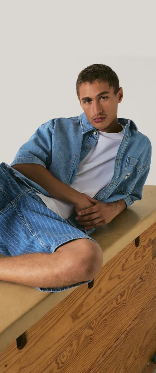 Junger Mann, der sich auf einer Holzoberfläche zurücklehnt, trägt ein blaues Jeanshemd und Shorts über einem weissen T-Shirt und schaut mit neutralem Ausdruck in die Kamera.