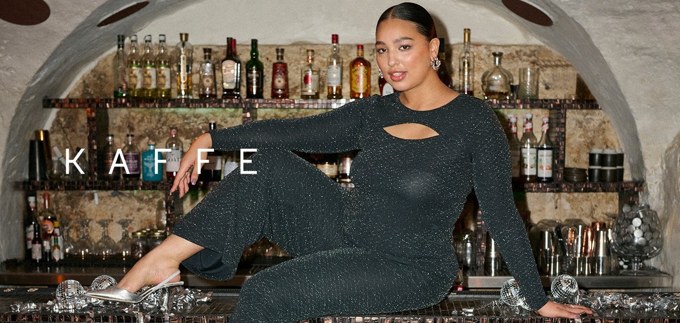 Černý třpytivý overal s dlouhými rukávy a výřezem, doplněný o stříbrné baleríny. Pozadí znázorňuje bar s lahvemi.