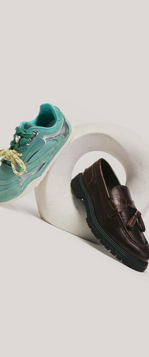 Ein türkisfarbener Sneaker mit gemischten Schnürsenkeln und ein dunkelbrauner Lederloafer auf einem weißen, gesprenkelten runden Podest positioniert.
