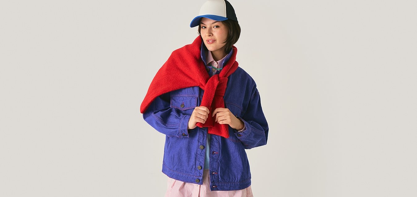 Mladá žena v modré bundě s červeným šátkem přehoděným přes ramena a bílou a modrou baseballovou čepicí stojí před jednobarevným pozadím.