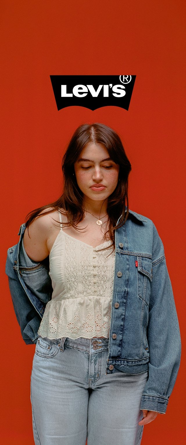 Frau trägt helle Jeans, eine teilweise schulterfreie Jeansjacke und ein weißes Spitzenoberteil, steht vor einem orangefarbenen Hintergrund mit dem Levi's-Logo oben.