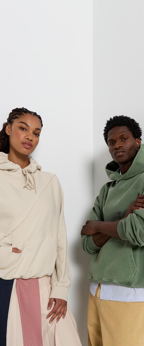 Zwei Personen stehen nebeneinander. Eine trägt einen hellbeigen Hoodie mit einem strukturierten diagonalen Design; die andere trägt einen sanften grünen Hoodie.