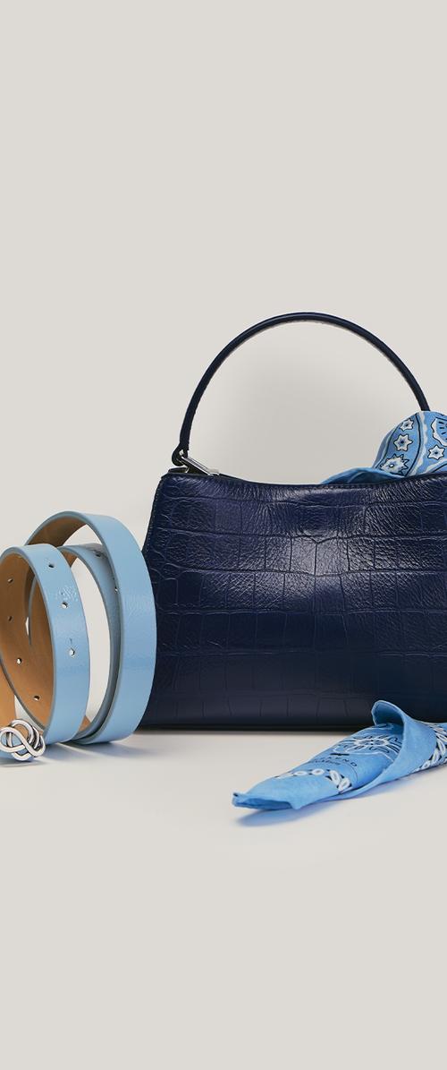 Bolso de mano texturizado azul marino con un asa, pañuelo azul con estampado doblado dentro y cinturón de cuero azul claro enrollado a su lado sobre un fondo neutro.