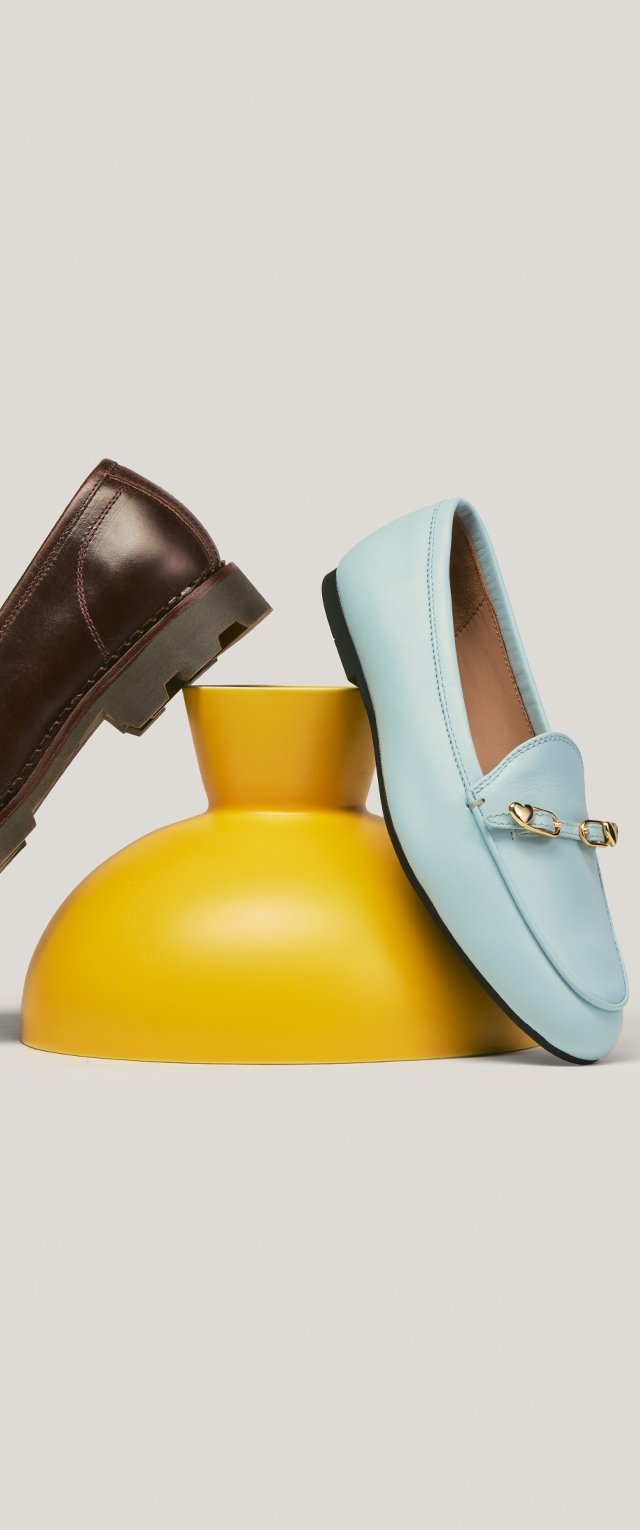 Barna bőrcsizma és világoskék mokaszin arany csattal, sárga váza tetején egy semleges háttér előtt.