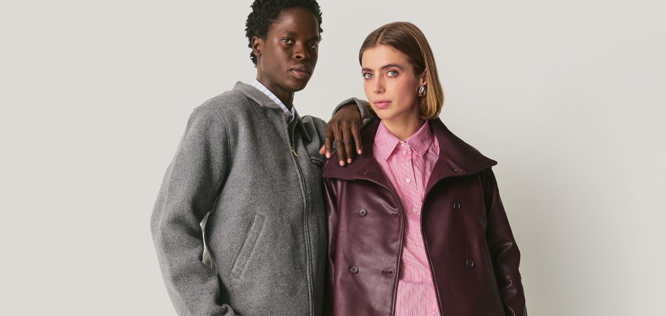 Dva lidé stojí blízko sebe; jeden má na sobě šedou bundu a kalhoty, druhý purpurovou koženou bundu, růžovou košili, zkrácené kalhoty a boty.