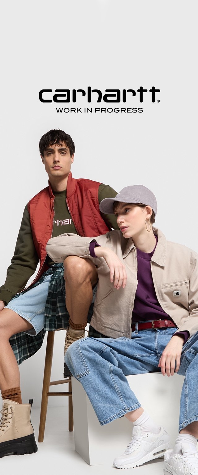 Due modelli posano, uno con un gilet rosso sopra una felpa verde e pantaloni di jeans corti, l'altro con una giacca beige e una maglietta viola, indossando cappelli e stivali.