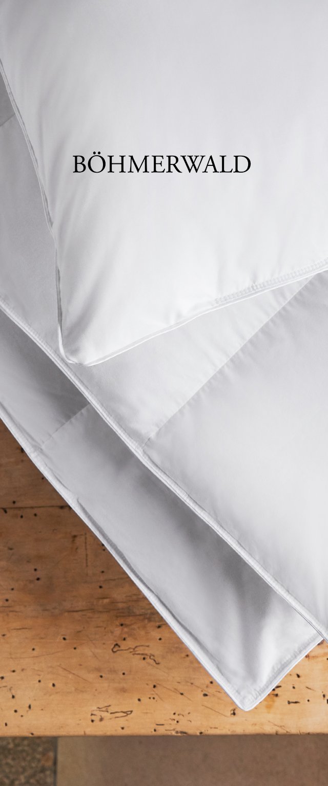 Weiße, gesteppten Kissen, die auf einer Holzoberfläche gestapelt sind, mit schwarzem "BÖHMERWALD"-Text auf dem obersten Kissen. Weicher Stoff und sauberes Design.