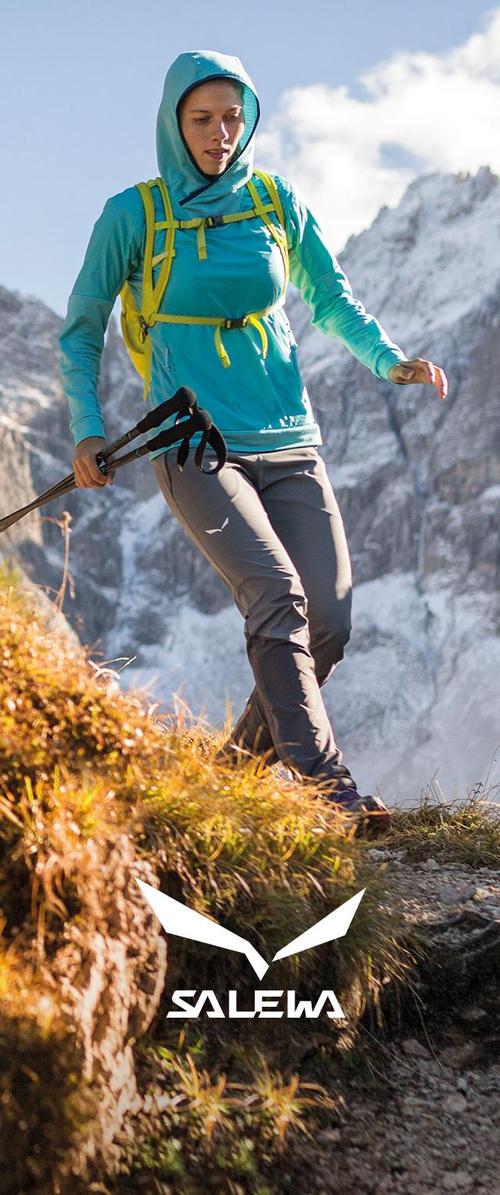 Turqouise hoodie met gele accenten, grijze wandelsbroek en trekkingstokken. Bergachtige achtergrond met rotsachtig terrein en gras.