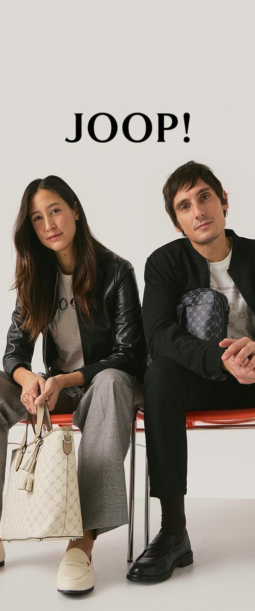 Dos personas sentadas llevan una chaqueta de cuero y una camisa estampada. Sostienen una bolsa beige y usan zapatos en crema y negro. Fondo sencillo.