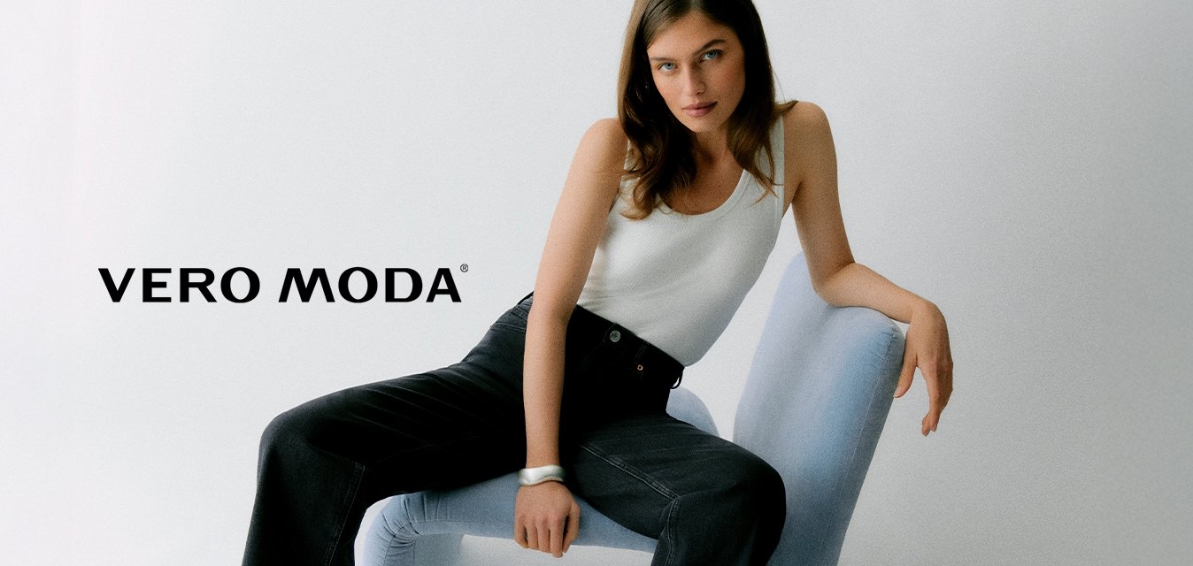 Žena s dlouhými hnědými vlasy sedí na světle modré moderní židli, má na sobě bílý tílko a černé kalhoty s širokými nohavicemi, na jednobarevném bílém pozadí.