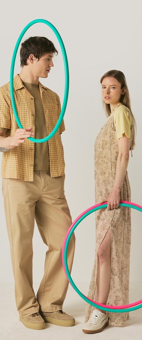 Mann im karierten Hemd hält einen blauen Hula-Hoop-Reifen vor sein Gesicht, Frau im geblümten Kleid hält einen rosa-blauen Hula-Hoop-Reifen an ihrer Seite.