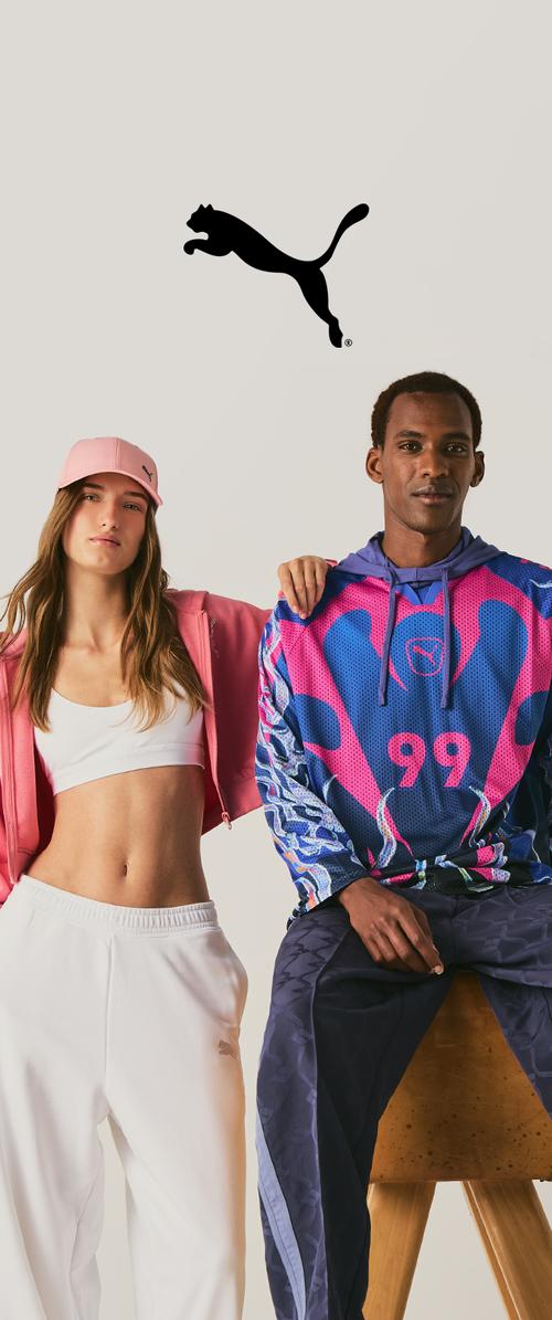 Junge Frau in pinker Sportbekleidung und Mann in buntem Hoodie und dunkler Hose posieren unter einem schwarzen springenden Puma-Logo auf schlichtem Hintergrund.