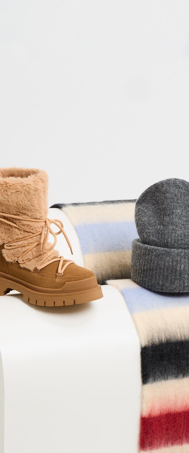 Stivaletti alla caviglia foderati in pelliccia marrone con dettagli in pizzo, abbinati a un berretto in maglia grigio e una coperta di lana a righe nei toni di blu, rosso e crema.