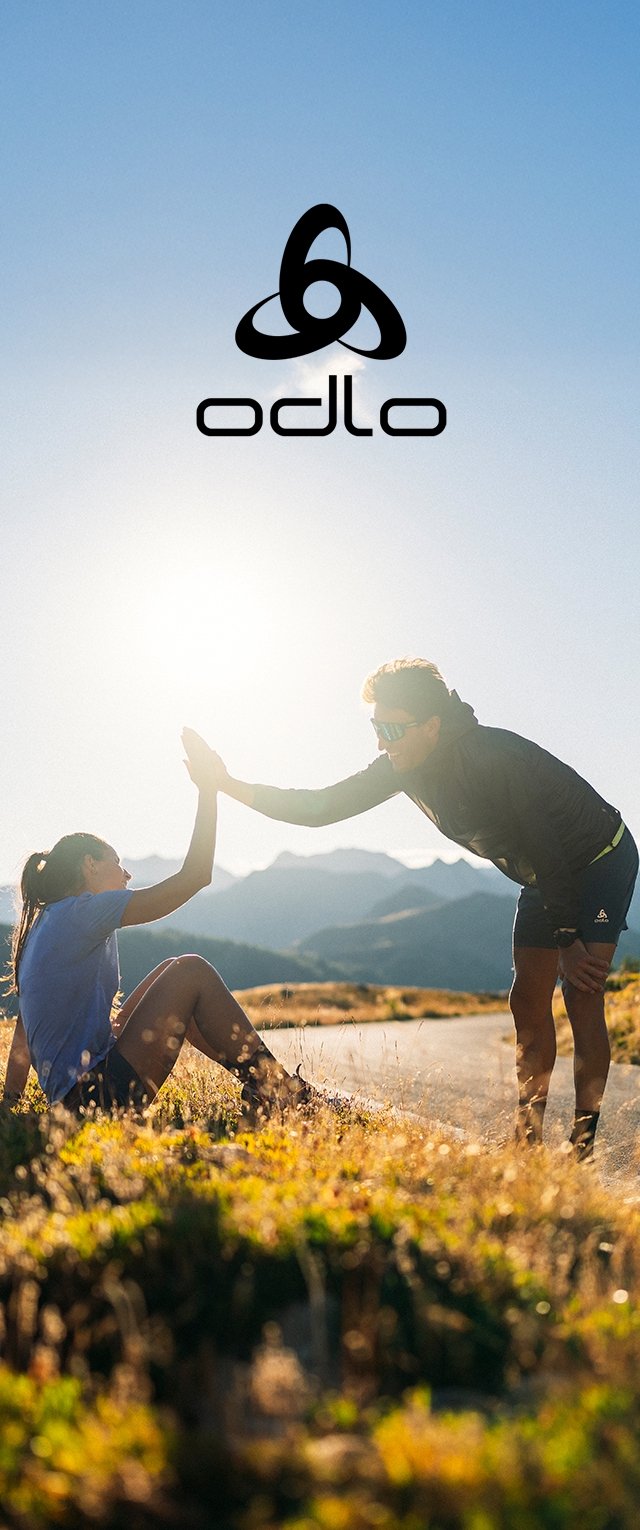 Zwei Personen interagieren im Freien in Sportbekleidung, wobei eine ihre Hand für ein High-Five hebt. Die Umgebung zeigt sonnenbeschienene Berge und Gras.