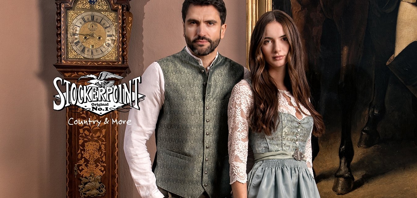 Muž a žena stojí uvnitř oblečeni v tradičním bavorském kroji, vedle starožitných dřevěných orloje a velkého rámovaného obrazu.